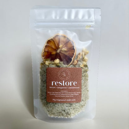 A clear resealable stand-up pouch containing a bath soak mixture with visible dried botanical pieces and a brown label reading 'restore lemon + bergamot + cedarwood'.