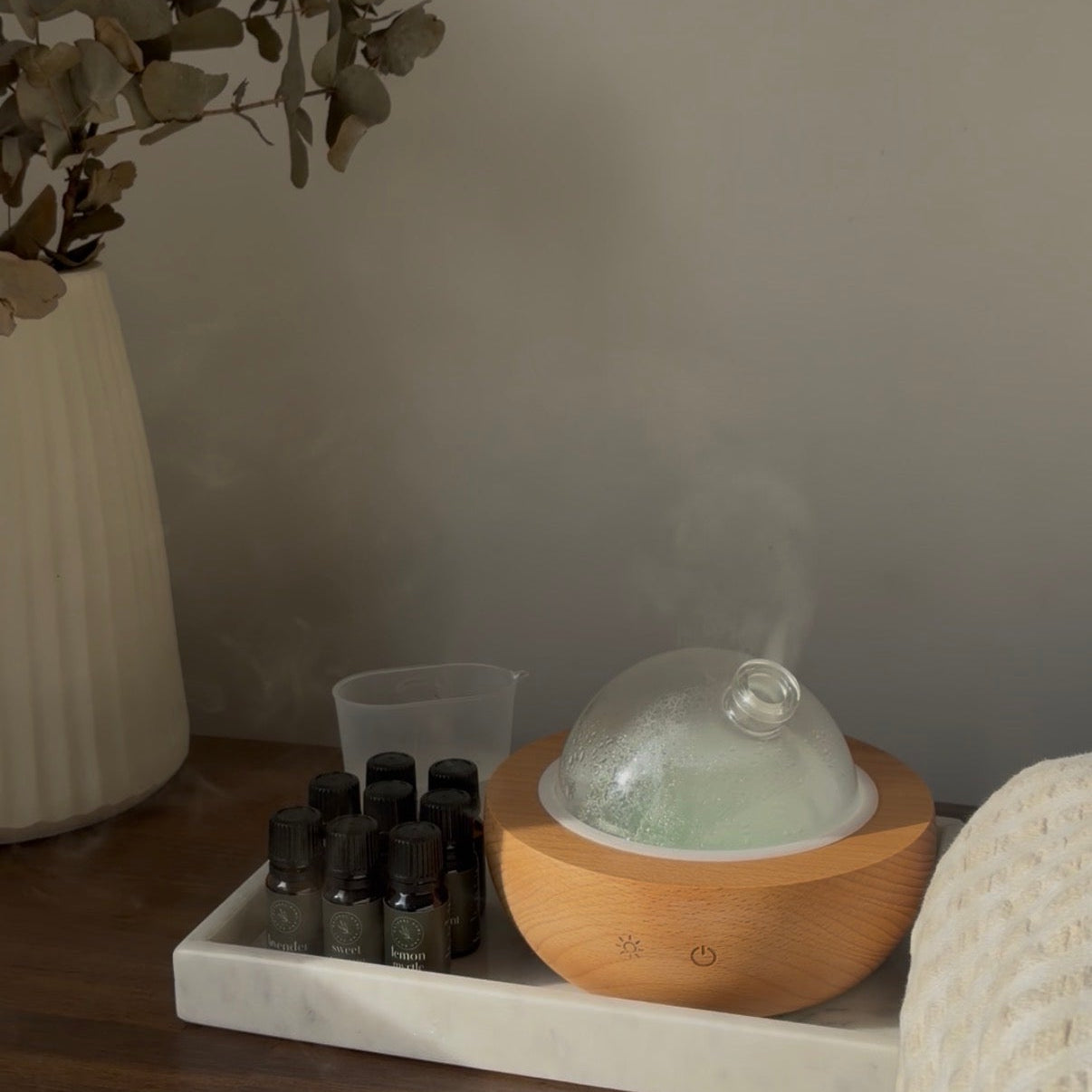 A wooden diffuser on a tray with essential oils and a vase of eucalyptus in a room setting.