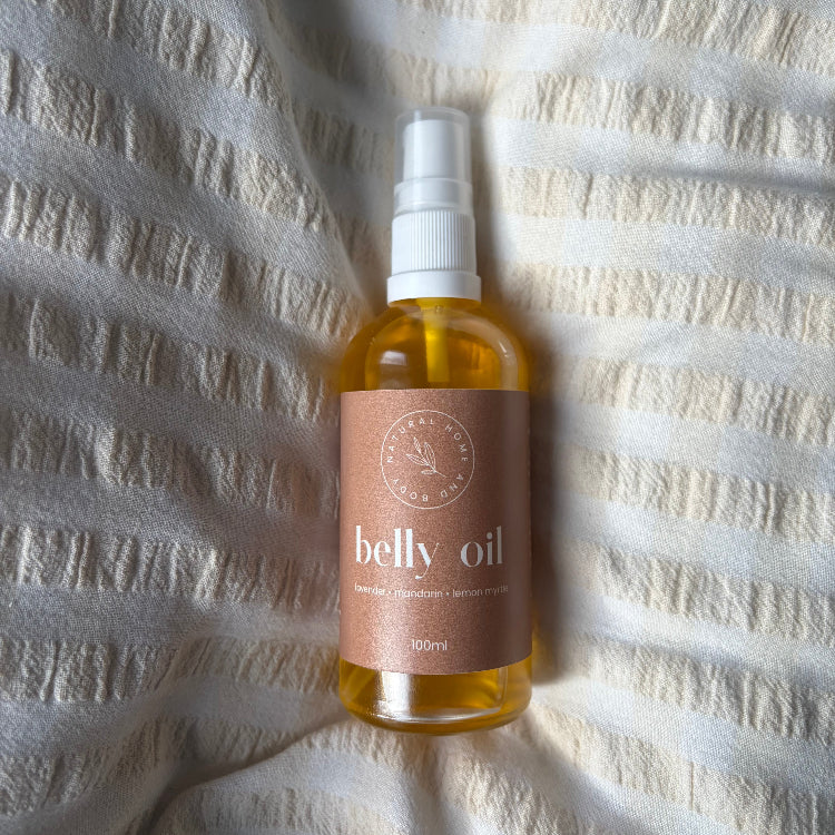 Belly oil in clear bottle with white serum pump, orange liquid and a burnt orange label. Centred on a beige bedsheet.